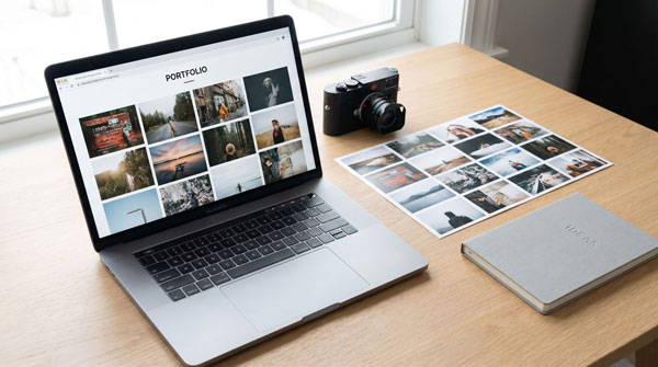 A photographer's desk with a laptop showing a responsive image gallery website, surrounded by a camera, printed photos in a grid layout, and a notebook, with soft natural lighting.